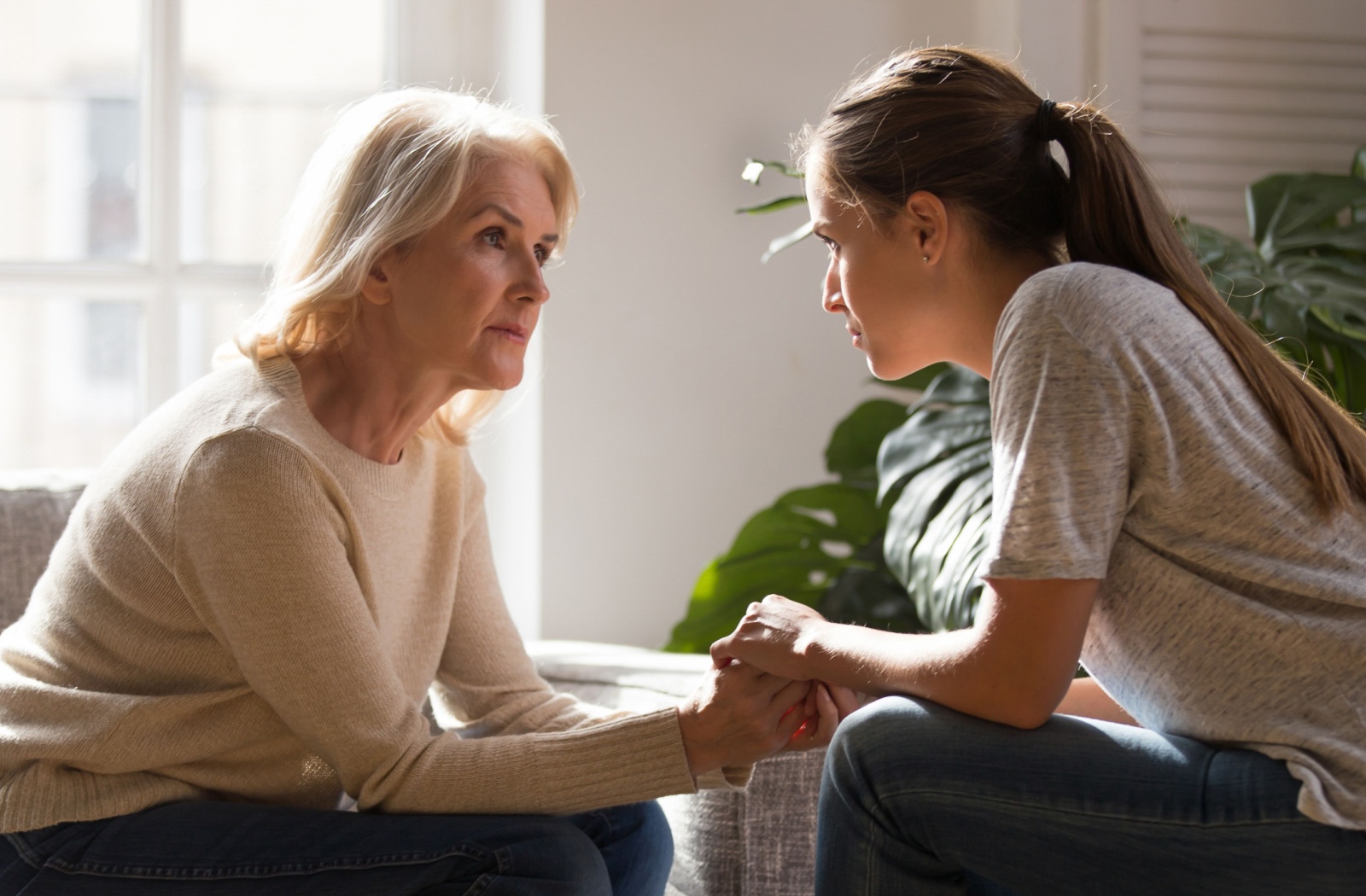 An adult child holding her mother's hands while sitting on the couch and having a discussion.