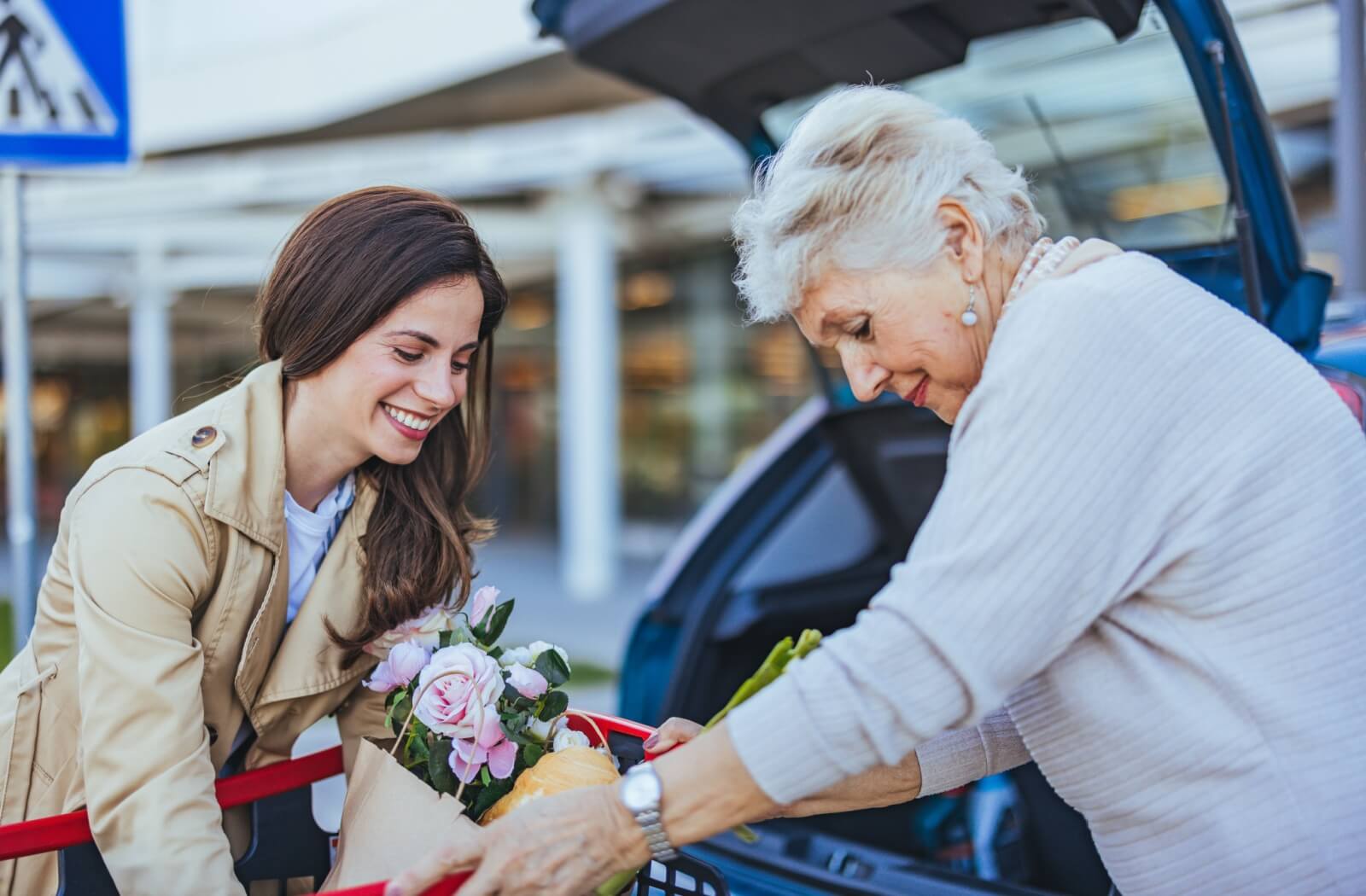 A smiling adult gives their senior parent a hand loading a bag into the back of their vehicle.