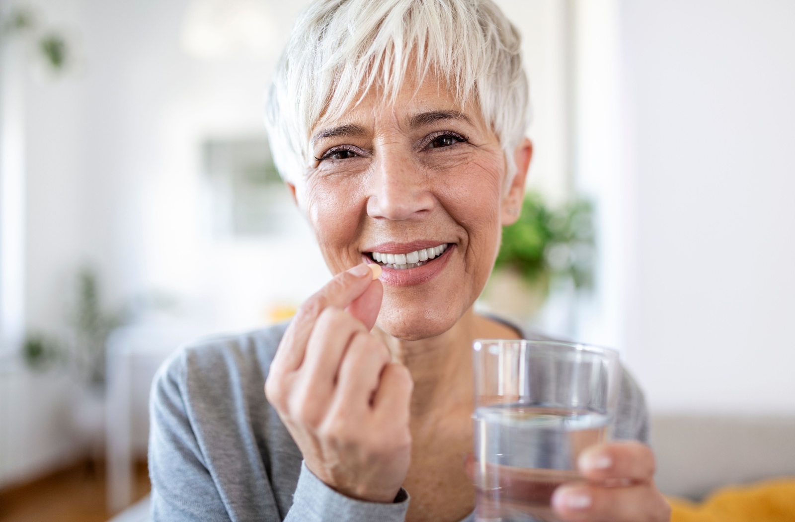 A smiling older adult holding a glass of water in one hand and a multivitamin pill in the other.