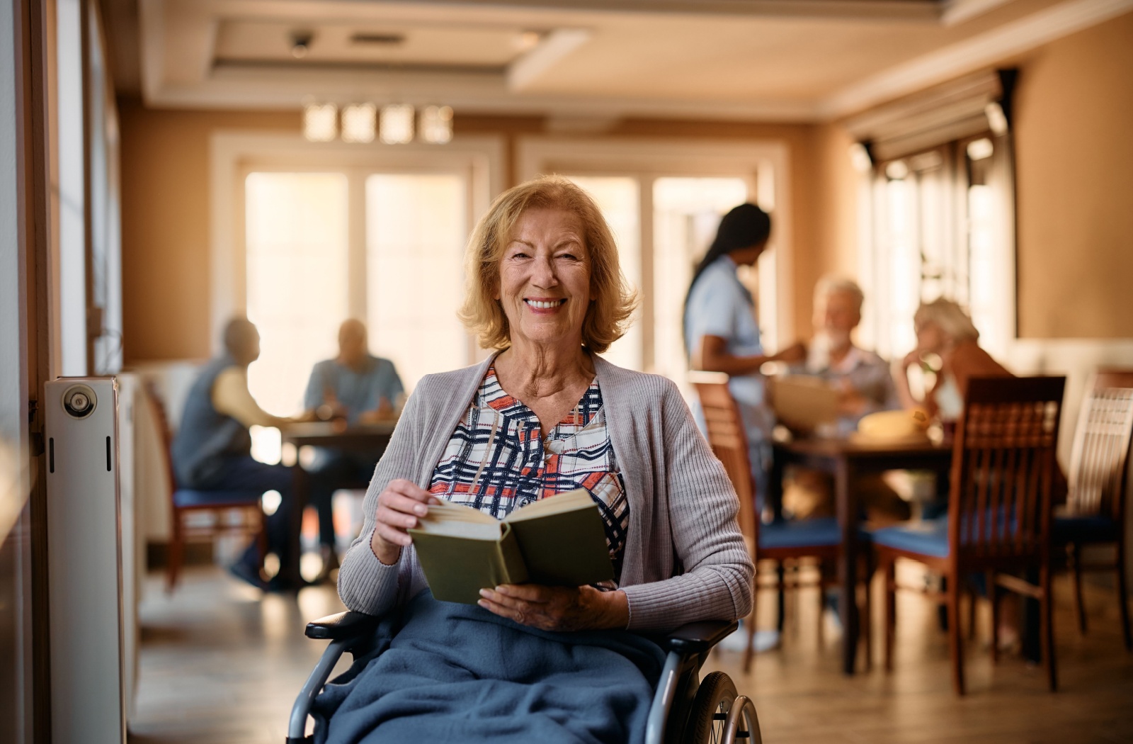 An older adult in the common area of assisted living smiling and reading their book.