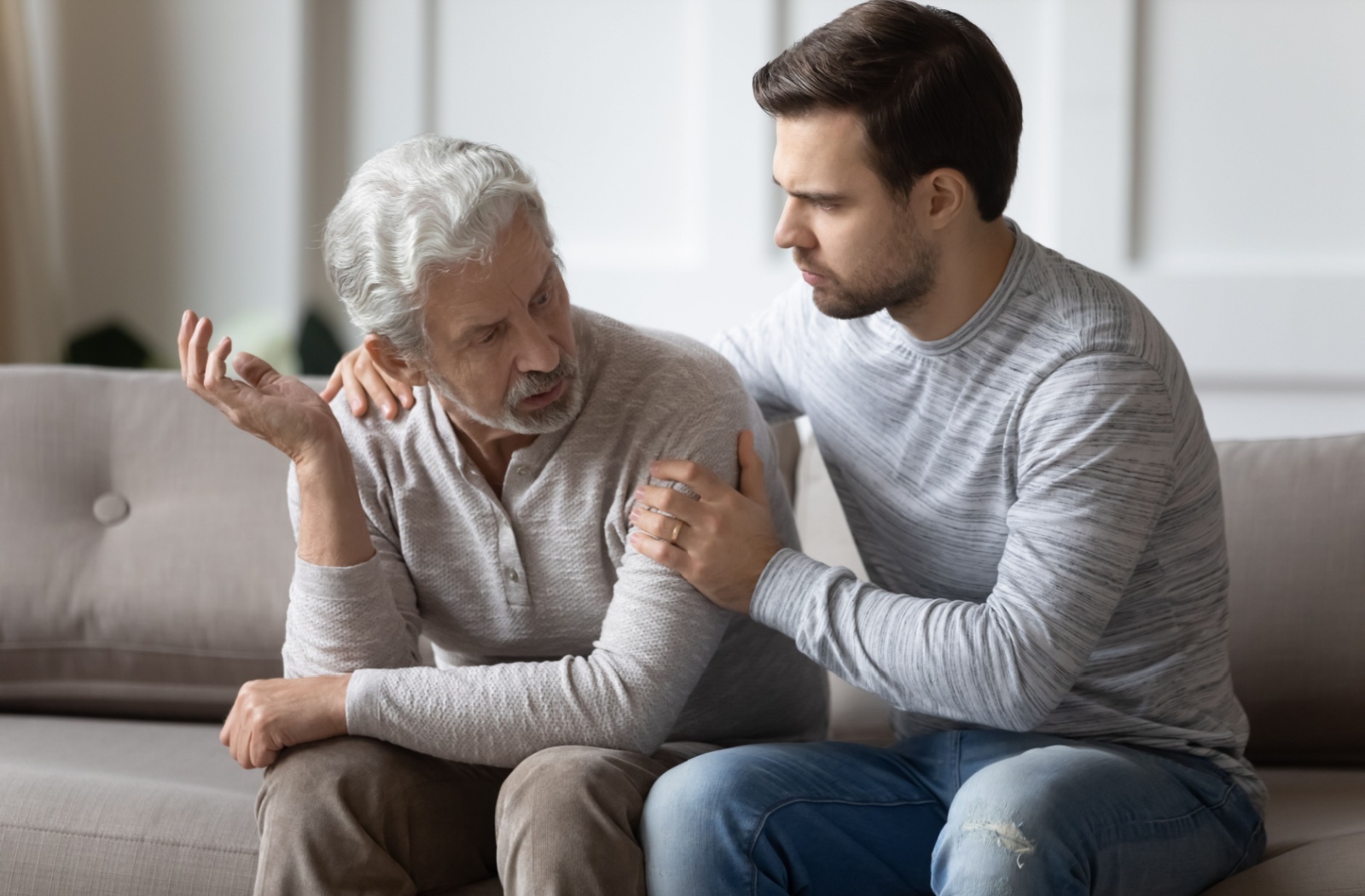 An adult child comforting their parent with Alzheimer's, sitting on the couch and talking together.