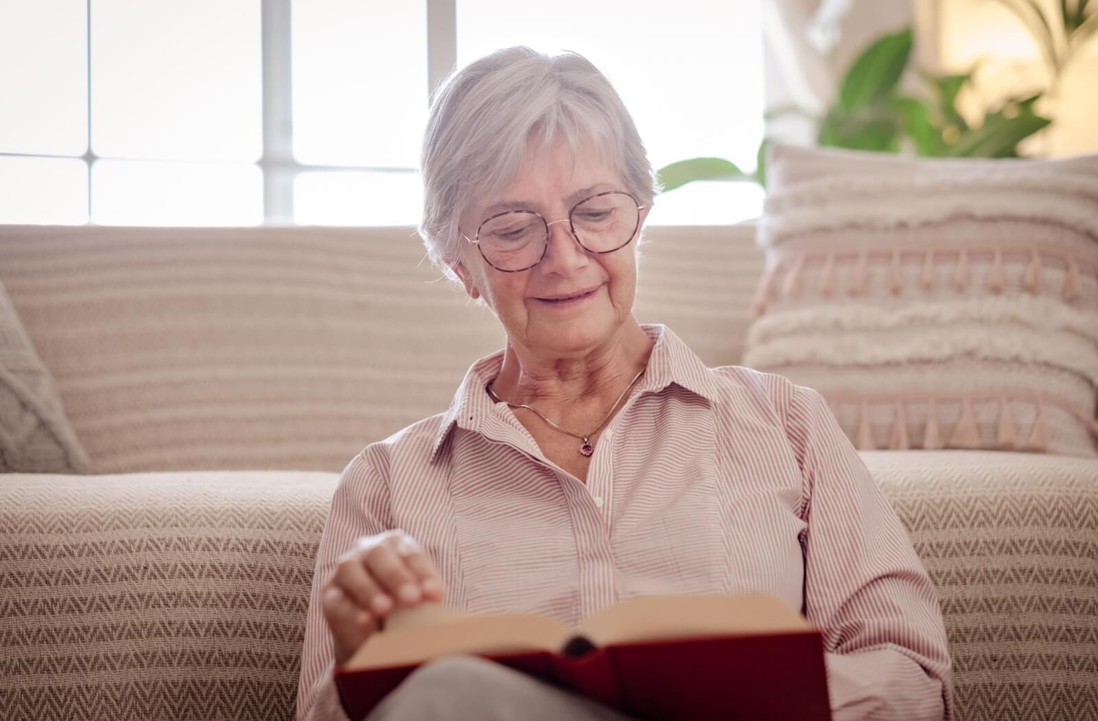 Older adult reading a book sitting against a cushioned couch with soft lighting and a calm focused expression
