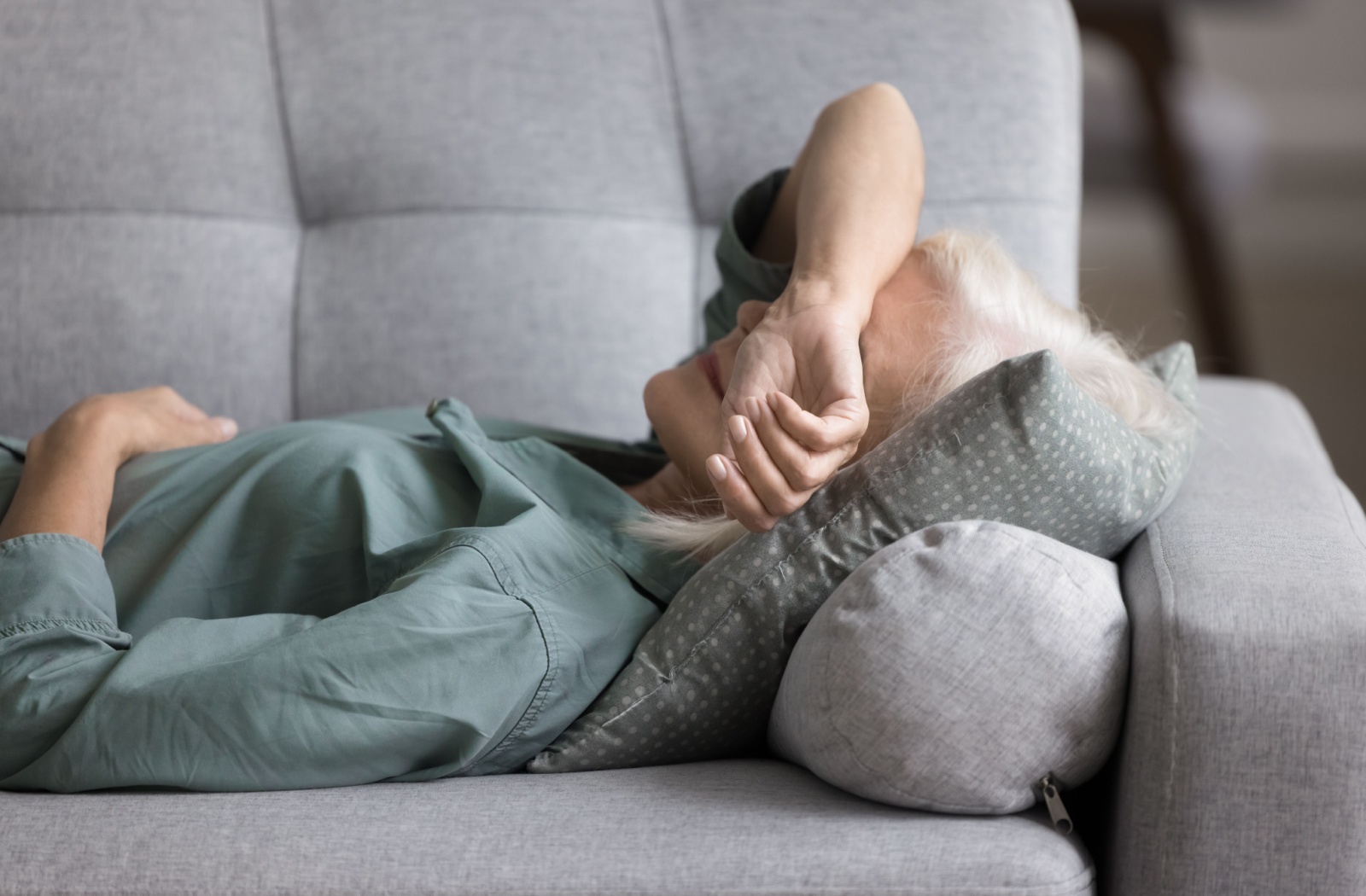 A senior lies on a sofa with their arm over their face, feeling fatigued and unmotivated.