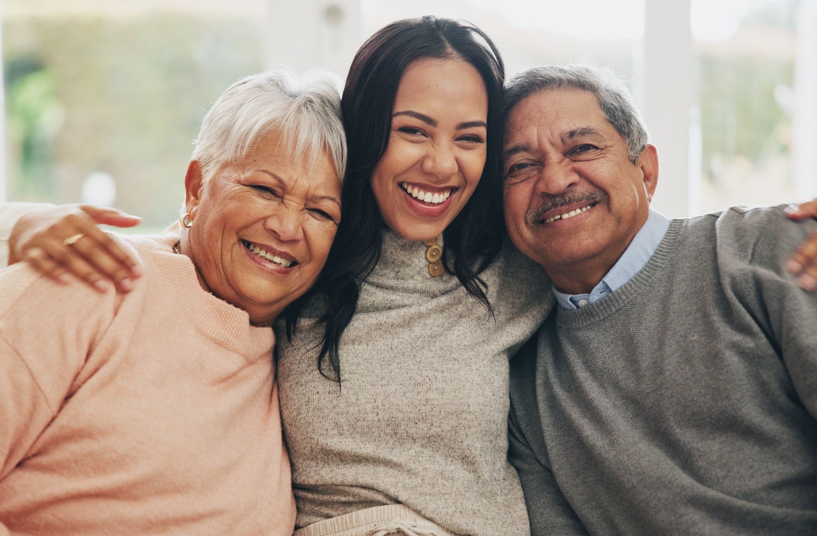 An adult child sits on a couch with their arms around their older parents.