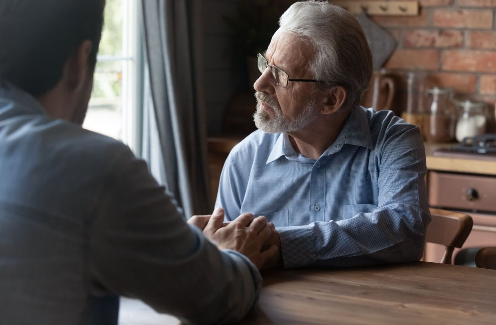 An adult child holds the hands of their older parent, who stares out of the window with a concerned expression.
