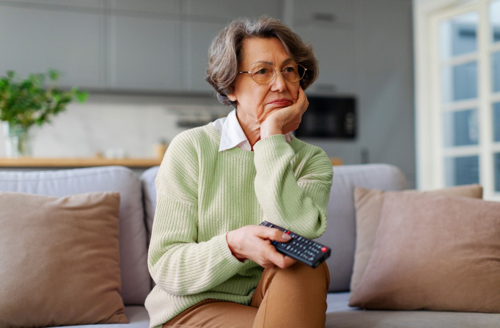 A senior sits on a couch holding a television remote while watching a movie.
