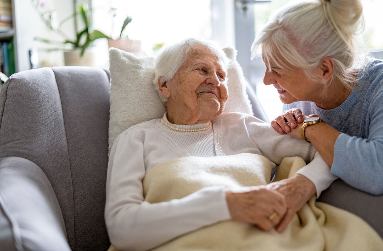 An adult child comforts their senior parent with dementia while they relax in a gray armchair.